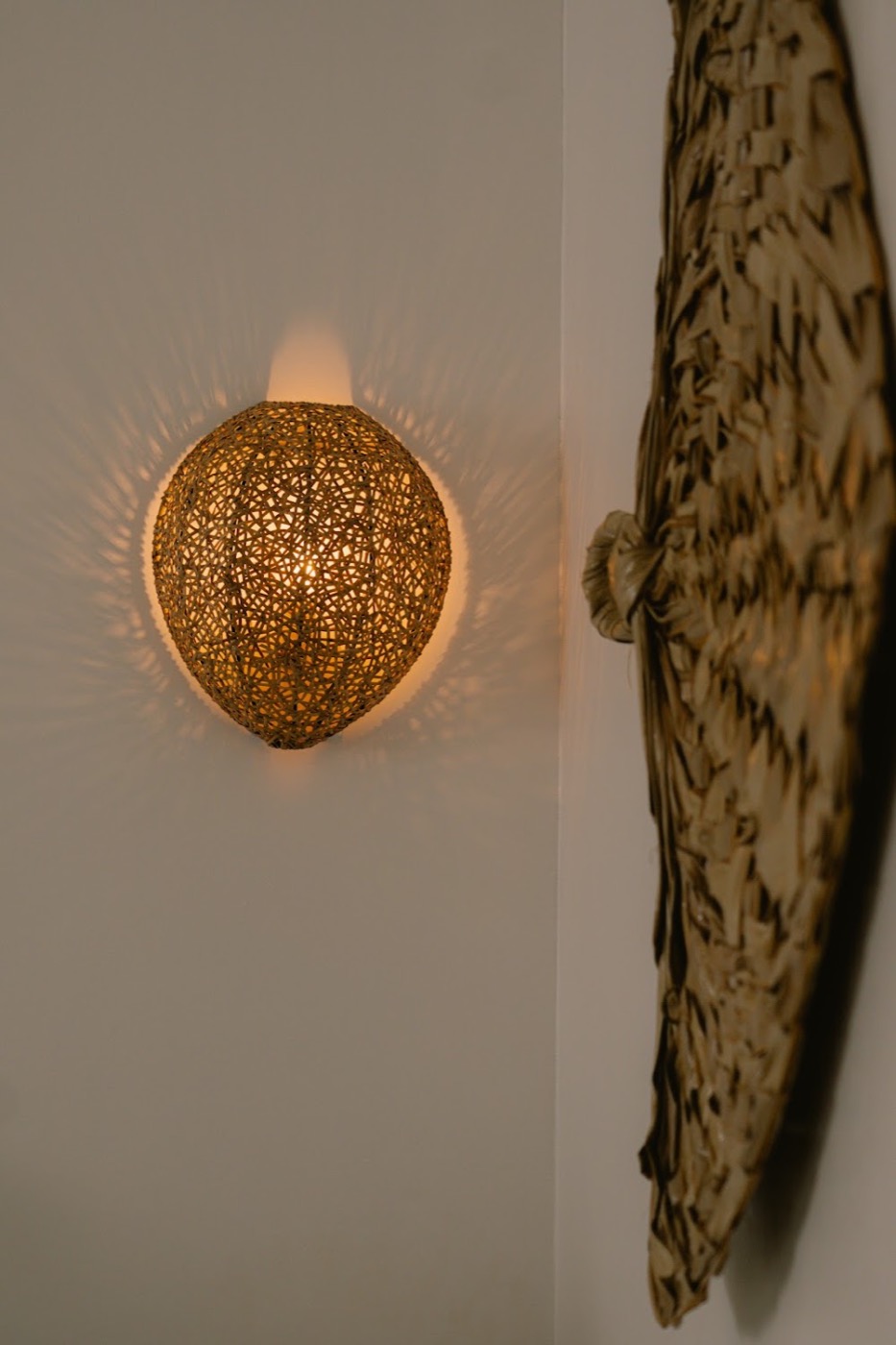 A glowing woven lampshade casting patterned light beside a textured native wall piece